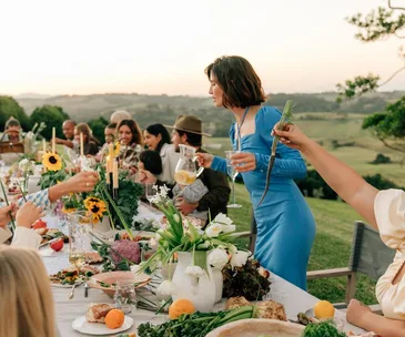 People enjoy an outdoor meal at a long table adorned with sunflowers and platters of food against a scenic background.