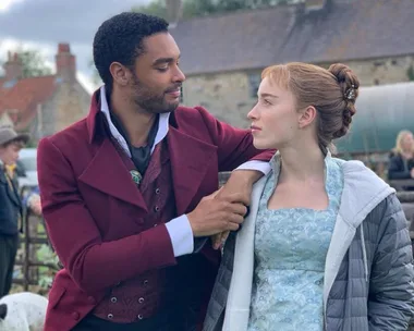 Regency couple in costume on set, man in burgundy coat, woman in blue dress, embracing and looking at each other.