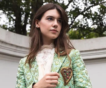 Young woman in a floral jacket with a heart brooch at the 2021 BAFTA red carpet event, outdoors with trees in background.