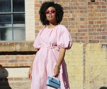 Person wearing a pink dress and sunglasses, holding a small blue purse, stands against a brick wall.