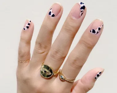 A hand with cow-print nail art and gold rings featuring an eye design on a light background.