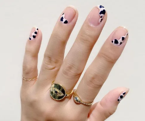 A hand with cow-print nail art and gold rings featuring an eye design on a light background.