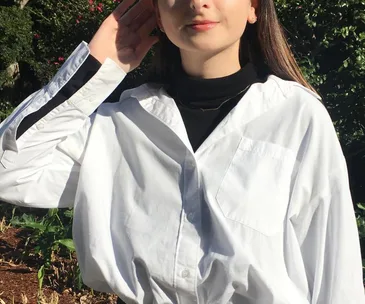 Woman wearing a white button-up shirt over a black turtleneck, standing outside in sunlight.