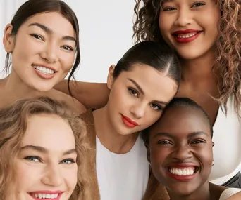 Five women smiling close together in a promotional image for Rare Beauty, featuring diverse models with bright lipstick.