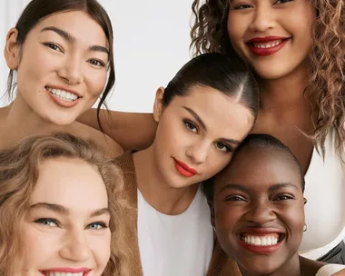 Five women smiling close together in a promotional image for Rare Beauty, featuring diverse models with bright lipstick.