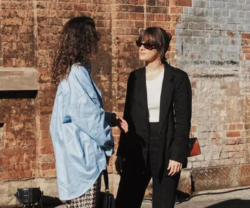 Two women wearing fashionable outfits converse outdoors against a brick wall. One wears sunglasses, the other a blue shirt.