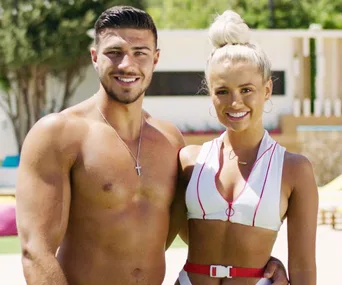 A smiling couple from Love Island UK 2021, one man shirtless, one woman in a white top with a bun hairstyle, outdoors.