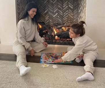 A woman and child in loungewear play a board game by the fireplace.