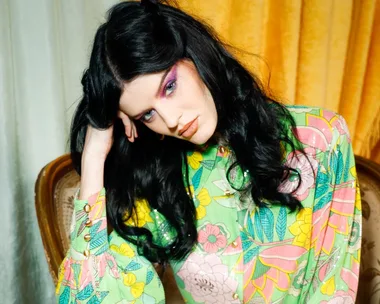 Woman in a colorful floral outfit with vibrant makeup and long dark hair sits on a vintage chair with a pensive expression.