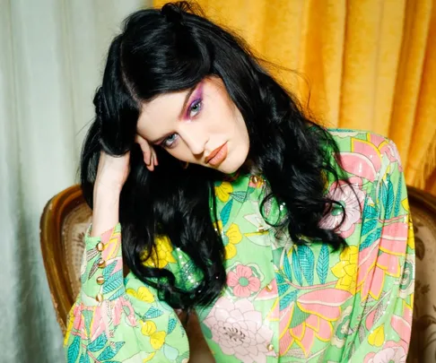 Woman in a colorful floral outfit with vibrant makeup and long dark hair sits on a vintage chair with a pensive expression.