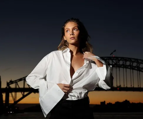 Model in white shirt poses at dusk in front of Sydney Harbour Bridge.