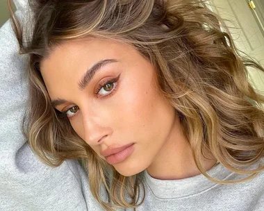 Close-up of a person with wavy, biscuit-brunette hair and natural makeup, wearing a light gray top.