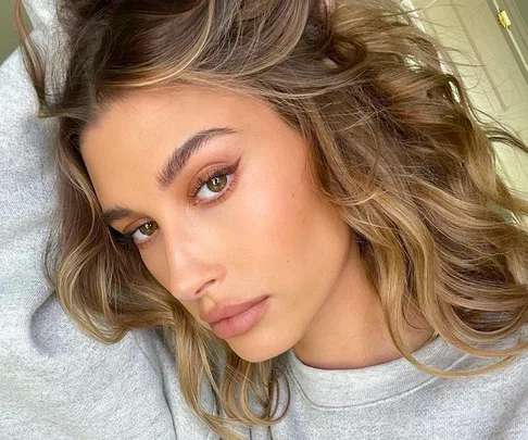 Close-up of a person with wavy, biscuit-brunette hair and natural makeup, wearing a light gray top.