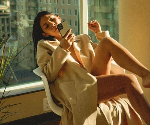 Person relaxing on a chair by a window, wearing a beige coat, looking at a phone.