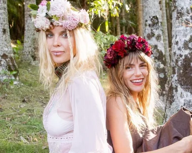 Two women seated back-to-back outdoors, wearing floral crowns and light dresses, smiling against a woodsy backdrop.