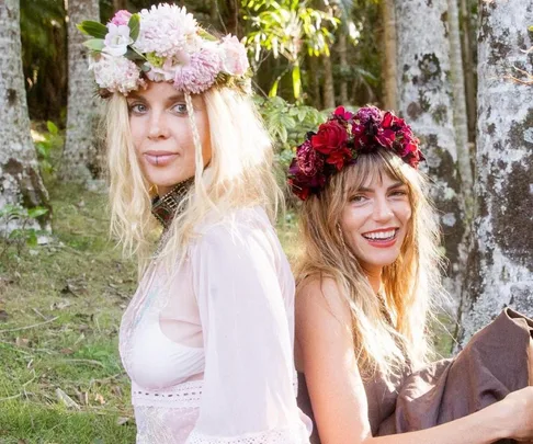 Two women seated back-to-back outdoors, wearing floral crowns and light dresses, smiling against a woodsy backdrop.