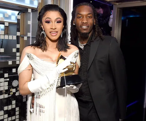 Woman in white gown holding a Grammy next to a man in a black suit, smiling at a glamorous event.