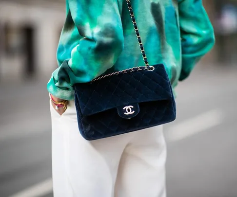 Woman in a green tie-dye shirt and white pants holds a blue Chanel quilted handbag with a chain strap.