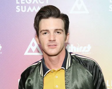 A man with dark hair in a black jacket and yellow shirt stands against a background with logos.