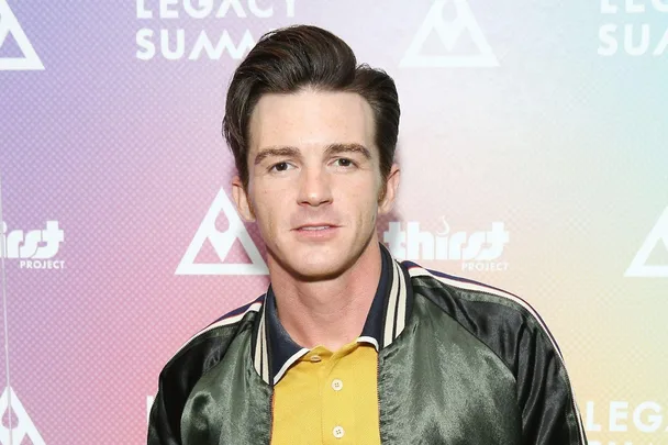 A man with dark hair in a black jacket and yellow shirt stands against a background with logos.