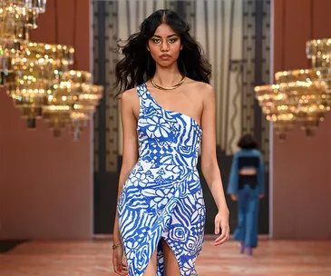 Model in a blue and white one-shoulder floral dress walks runway at Australian Fashion Week 2021 amidst chandeliers.