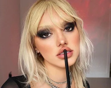 Woman with blonde hair and heavy dark eye makeup, holding a brush to her lips, wearing a black top and chain necklace.