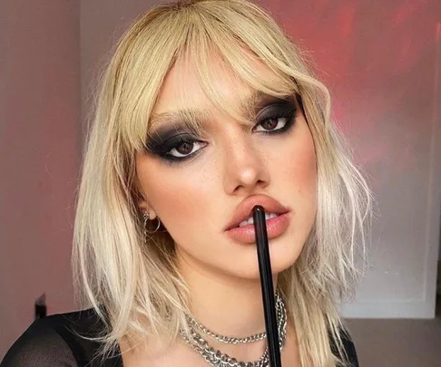 Woman with blonde hair and heavy dark eye makeup, holding a brush to her lips, wearing a black top and chain necklace.