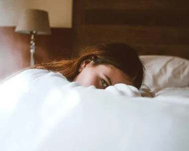 Woman lying in bed, partially covered by a blanket, with a soft lamp in the background.