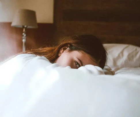 Woman lying in bed, partially covered by a blanket, with a soft lamp in the background.