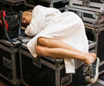 Woman in a white robe and sneakers sleeping on stacked equipment cases backstage.