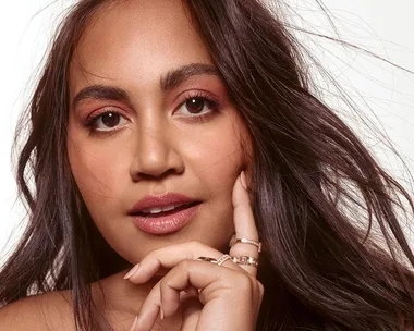 A close-up image of Jessica Mauboy with long wavy hair, wearing natural makeup, and gently holding her chin with one hand.