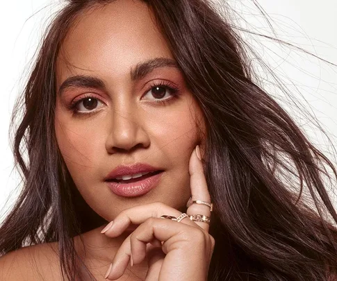 A close-up image of Jessica Mauboy with long wavy hair, wearing natural makeup, and gently holding her chin with one hand.