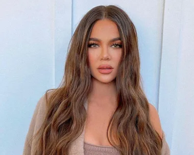 A person with long, wavy brown hair, wearing a beige outfit, against a light background.