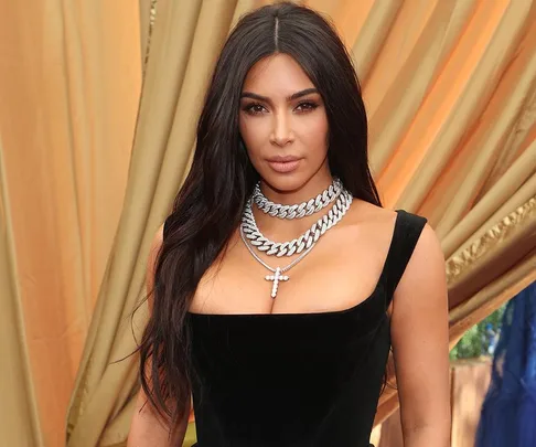 Woman in a black dress with silver layered necklaces stands in front of a gold curtain.