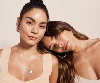 Two women pose closely together, wearing simple jewelry and neutral tops against a beige background.