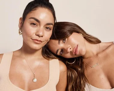 Two women pose closely together, wearing simple jewelry and neutral tops against a beige background.