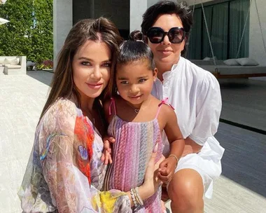 Three people posing happily outdoors, including a child in a colorful dress, with modern patio furniture in the background.