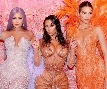 Women in elegant dresses pose in front of a wall of pink roses, showcasing fashion at a high-profile event.