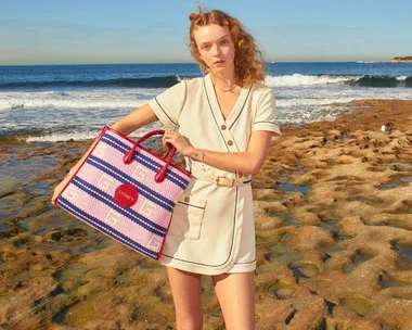 A person in a cream outfit holds a colorful Gucci bag at the beach, with waves and rocks in the background.