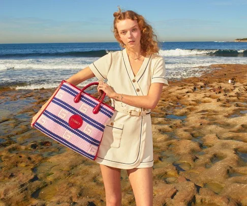 A person in a cream outfit holds a colorful Gucci bag at the beach, with waves and rocks in the background.