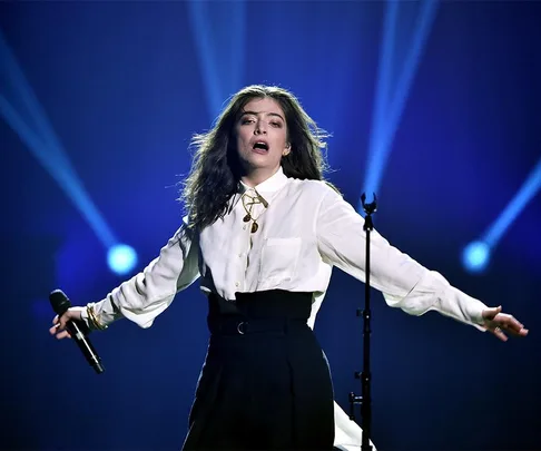 Singer performing on stage, wearing a white blouse and black pants, with blue spotlights in the background.