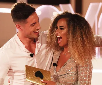 Two Love Island UK contestants celebrate holding a gold heart envelope, both wearing microphones.