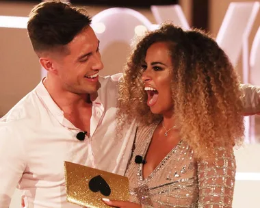 Two Love Island UK contestants celebrate holding a gold heart envelope, both wearing microphones.