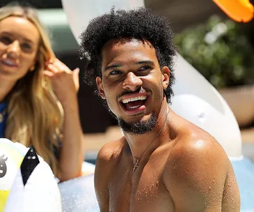 Man smiling in a swimming pool, with another person blurred in the background.