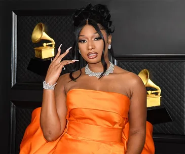 Megan Thee Stallion in an orange strapless gown at the Grammy Awards, with diamond jewelry and Grammys in the background.