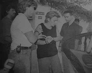 Black and white image shows a woman being escorted by police officers to a car.