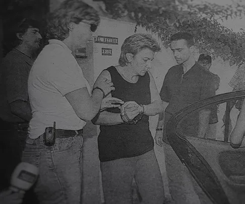 Black and white image shows a woman being escorted by police officers to a car.