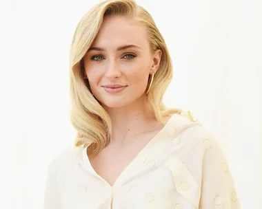 Blonde woman with long wavy hair, wearing a dotted cream blouse, smiling softly against a white background.