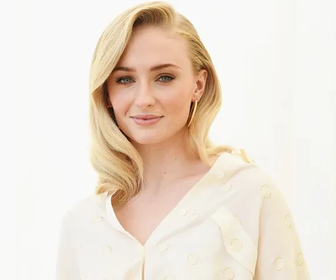 Blonde woman with long wavy hair, wearing a dotted cream blouse, smiling softly against a white background.