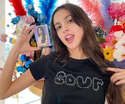 Young woman in a "SOUR" shirt holds a cassette tape, surrounded by colorful flowers and feathers.